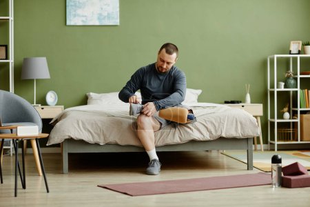 Foto de Retrato de cuerpo entero del hombre adulto arreglando la pierna protésica y preparándose para hacer ejercicio en casa, espacio para copiar - Imagen libre de derechos