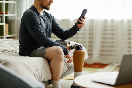 Téléchargez les photos : Portrait latéral de l'homme avec prothèse de jambe à l'aide d'un smartphone assis sur le lit à la maison - en image libre de droit