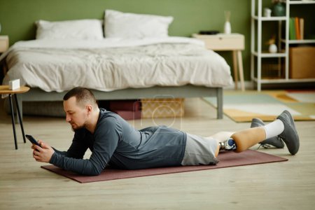 Téléchargez les photos : Portrait latéral de l'homme avec une jambe prothétique posée sur le sol à la maison et utilisant un smartphone - en image libre de droit