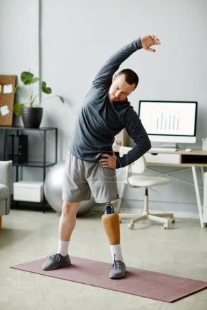 Foto de Retrato vertical de cuerpo entero de hombre con pierna protésica estirándose en casa de pie sobre esterilla de yoga - Imagen libre de derechos