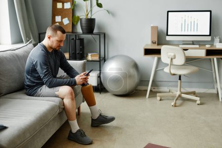 Téléchargez les photos : Portrait latéral de l'homme adulte avec prothèse de jambe à l'aide d'un smartphone tout en étant assis sur le canapé dans un espace minimal à la maison, copier - en image libre de droit