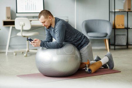 Foto de Retrato de vista lateral de hombre adulto con prótesis de pierna haciendo ejercicio con pelota de fitness en casa y usando smartphone - Imagen libre de derechos