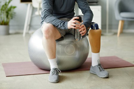 Téléchargez les photos : Gros plan de section basse de l'homme avec la jambe prothétique assis sur la boule de forme physique et tenant la bouteille d'eau après l'entraînement à la maison - en image libre de droit