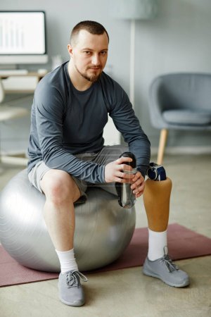 Téléchargez les photos : Portrait vertical complet de l'homme avec une jambe prothétique assise sur une balle de fitness et tenant une bouteille d'eau après l'entraînement à la maison - en image libre de droit
