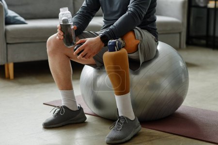 Téléchargez les photos : Gros plan d'un homme méconnaissable avec une jambe prothétique assise sur une balle de fitness et tenant une bouteille d'eau après l'entraînement à la maison - en image libre de droit