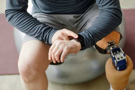 Téléchargez les photos : Gros plan de l'homme avec prothèse jambe vérifier smartwatch assis sur la balle de remise en forme pendant l'entraînement à la maison - en image libre de droit