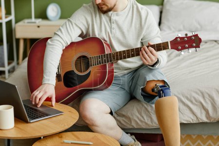 Foto de Primer plano del hombre con la pierna protésica tocando la guitarra en casa y utilizando el ordenador portátil, espacio de copia - Imagen libre de derechos