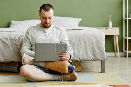 Photo for Full length portrait of man with prosthetic leg using laptop while sitting on floor at home, copy space - Royalty Free Image
