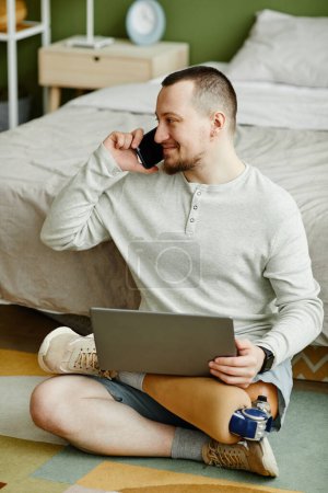 Téléchargez les photos : Portrait complet d'un homme souriant avec une jambe prothétique à l'aide d'un ordinateur portable assis sur le sol à la maison - en image libre de droit