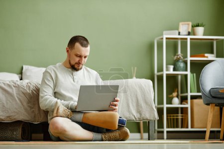 Foto de Retrato mínimo del hombre con la pierna protésica usando el ordenador portátil mientras está sentado en el suelo en casa, espacio de copia - Imagen libre de derechos