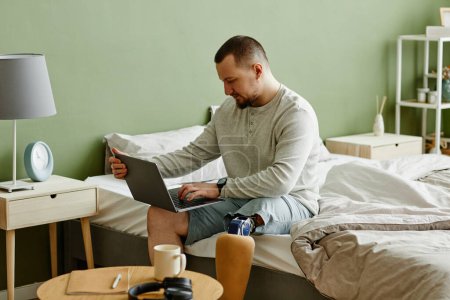 Photo for Portrait of man with prosthetic leg using laptop while sitting on bed at home, copy space - Royalty Free Image