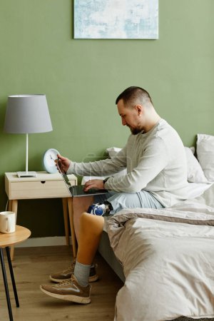 Photo for Side view portrait of man with prosthetic leg using laptop while sitting on bed at home - Royalty Free Image