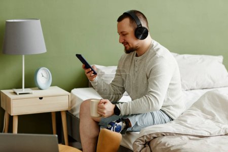 Téléchargez les photos : Portrait latéral d'un homme souriant avec une jambe prothétique profitant du matin à la maison et écoutant de la musique avec écouteurs, espace de copie - en image libre de droit