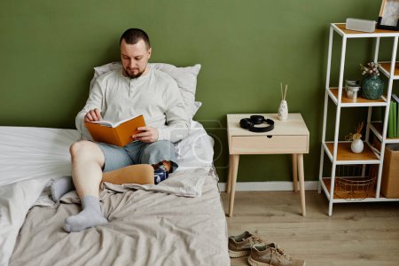 Photo for High angle view at young man with prosthetic leg reading book in bed while relaxing at home, copy space - Royalty Free Image