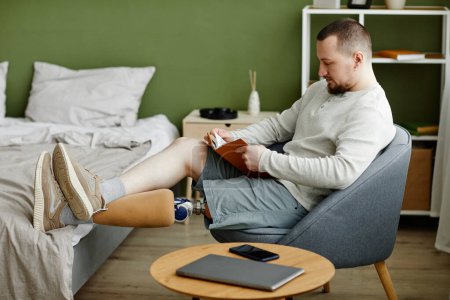 Téléchargez les photos : Portrait latéral de l'homme avec livre de lecture de jambe prothétique à la maison et relaxant, espace de copie - en image libre de droit