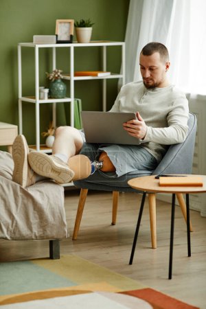 Téléchargez les photos : Portrait vertical complet de l'homme adulte avec prothèse de jambe relaxant à la maison et à l'aide d'un ordinateur portable - en image libre de droit