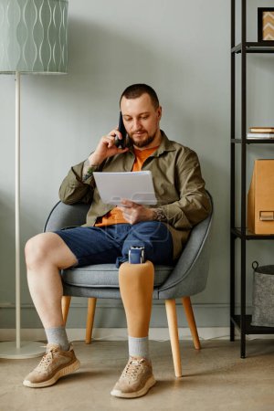 Téléchargez les photos : Portrait complet de l'homme avec la jambe prothétique appelant par téléphone tout en étant assis dans la chaise à l'intérieur de la maison - en image libre de droit