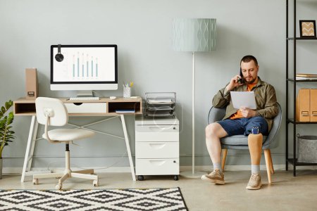 Photo for Full length portrait of man with prosthetic leg calling by phone while sitting in chair in home office interior, copy space - Royalty Free Image