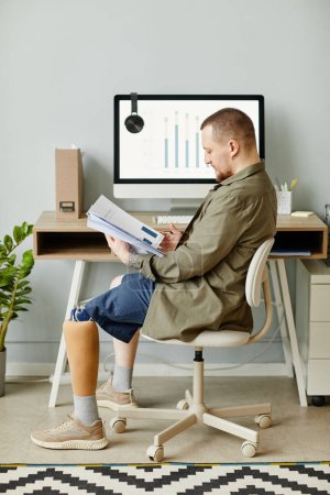Photo for Vertical full length portrait of man with prosthetic leg working at home office and reading documents - Royalty Free Image