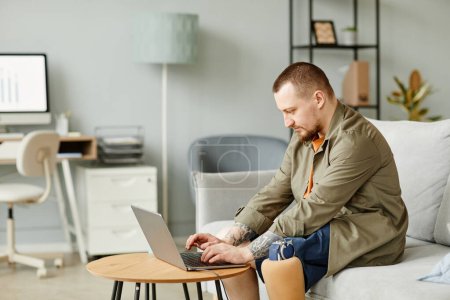 Téléchargez les photos : Portrait latéral de l'homme avec la jambe prothétique en utilisant un ordinateur portable tout en travaillant dans un intérieur de bureau minimal, espace de copie - en image libre de droit