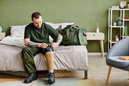 Foto de Retrato completo de veterano militar con pierna protésica que se pone uniforme del ejército en el dormitorio, espacio de copia - Imagen libre de derechos