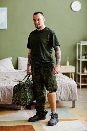 Téléchargez les photos : Portrait vertical d'un vétéran militaire avec une jambe prothétique portant l'uniforme de l'armée à la maison et regardant la caméra - en image libre de droit