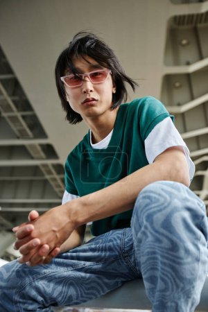 Téléchargez les photos : Portrait vertical de style de rue asiatique homme regardant la caméra à l'extérieur et portant des lunettes de soleil roses - en image libre de droit