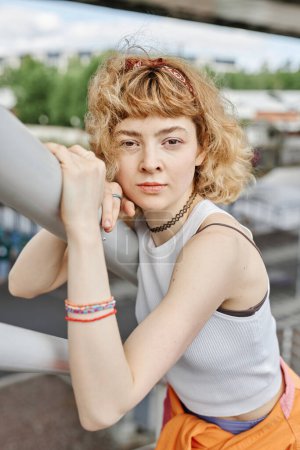 Téléchargez les photos : Portrait franc de jeune femme aux cheveux bouclés regardant la caméra à l'extérieur et portant une tenue tendance - en image libre de droit