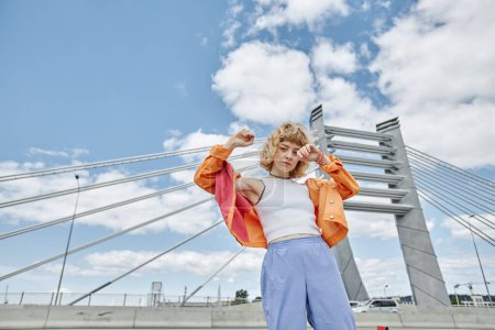Téléchargez les photos : Portrait taille haute de jeune femme libre portant des vêtements colorés dansant sur le pont de la ville et regardant la caméra contre le ciel, copier l'espace - en image libre de droit