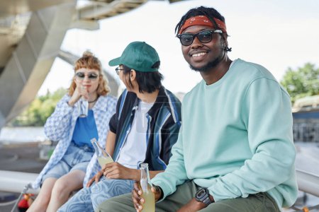 Téléchargez les photos : Un groupe diversifié de trois jeunes gens qui traînent ensemble en milieu urbain se concentre sur un homme noir souriant regardant la caméra - en image libre de droit