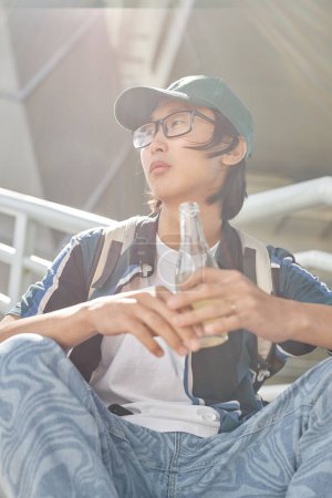 Photo for Vertical portrait of young Asian man wearing streetstyle clothes outdoors in sunlight and holding glass bottle - Royalty Free Image