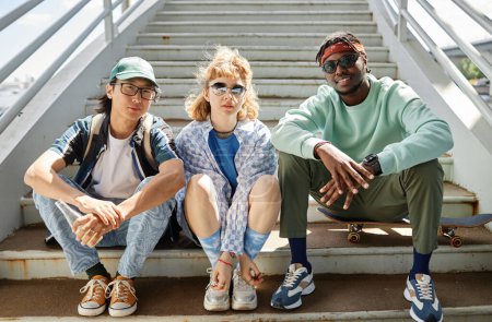 Téléchargez les photos : Groupe diversifié de jeunes gens portant des vêtements de style de rue à l'extérieur assis sur des escaliers en zone urbaine et regardant la caméra - en image libre de droit