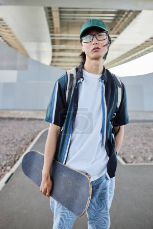 Foto de Retrato vertical del joven asiático con monopatín posando al aire libre en un entorno urbano - Imagen libre de derechos