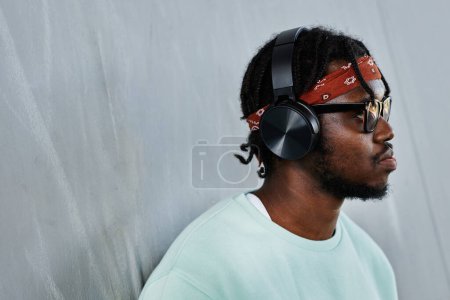 Foto de Retrato de vista lateral mínimo del joven hombre negro que usa auriculares junto a la pared de hormigón al aire libre, espacio para copiar - Imagen libre de derechos