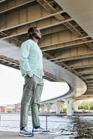 Photo for Full length side view of young black man wearing headphones in urban city setting outdoors - Royalty Free Image