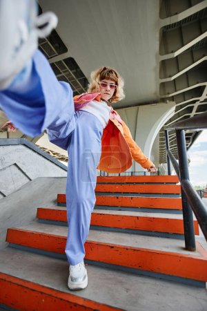 Téléchargez les photos : Portrait de jeune femme insouciante portant la mode de style de rue à l'extérieur en milieu urbain et donnant des coups de pied à la caméra - en image libre de droit
