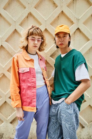 Téléchargez les photos : Portrait vertical de deux jeunes gens portant la mode colorée de style de rue posant contre le mur texturé à l'extérieur - en image libre de droit