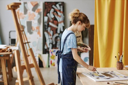 Foto de Vista lateral de la joven rubia en delantal de mezclilla pintando obras de arte con pinturas acrílicas mientras se inclina sobre el lugar de trabajo con lienzo - Imagen libre de derechos