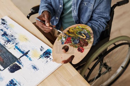 Téléchargez les photos : Vue ci-dessus du jeune homme noir avec palette de couleurs mélangeant peintures acryliques tout en étant assis sur fauteuil roulant par table en bois avec toile - en image libre de droit
