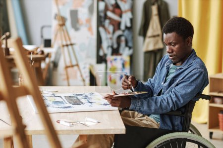 Téléchargez les photos : Jeune artisan afro-américain en fauteuil roulant mélangeant les couleurs sur la palette tout en étant assis à table en studio d'art et de peinture sur toile - en image libre de droit