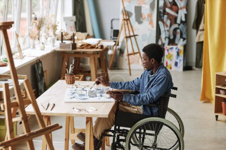 Photo for Young contemporary artist in wheelchair working over new painting while sitting in studio of arts, mixing paints and applying them on canvas - Royalty Free Image