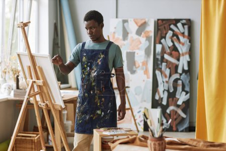 Téléchargez les photos : Jeune homme afro-américain sérieux en tablier de denim debout devant la toile sur chevalet et créer une nouvelle peinture à lesiure - en image libre de droit