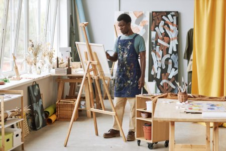 Téléchargez les photos : Jeune artiste masculin créatif sérieux dans le tablier debout devant le chevalet avec des œuvres d'art inachevées tout en travaillant sur une nouvelle peinture en studio - en image libre de droit