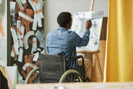 Photo for Rear view of African American male painter in wheelchair painting on canvas while sitting in front of easel in studio or classroom - Royalty Free Image