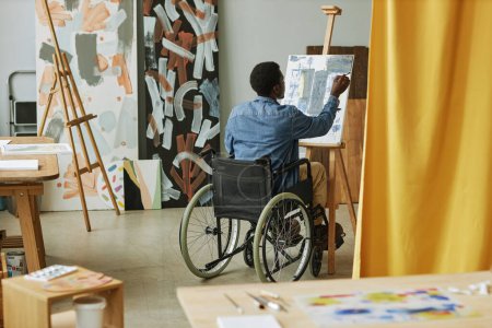 Téléchargez les photos : Vue arrière du jeune homme noir assis en fauteuil roulant devant le chevalet et peinture sur toile avec peintures acryliques en studio d'arts - en image libre de droit
