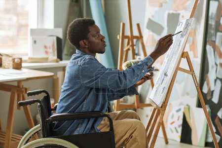 Photo for Young man with disability painting masterpiece on canvas with acrylic paints while sitting in wheelchair in front of easel in studio - Royalty Free Image