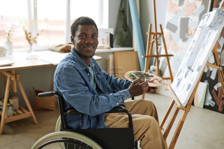 Foto de Joven pintor exitoso con discapacidad sentado en silla de ruedas delante de la cámara y mirándote mientras trabajas sobre una pintura nueva - Imagen libre de derechos