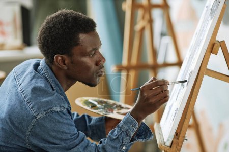 Téléchargez les photos : Vue latérale du jeune homme noir sérieux en denim chemise peignant de nouvelles œuvres d'art avec des peintures acryliques tout en étant assis devant le chevalet avec toile - en image libre de droit