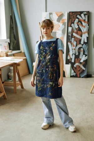 Photo for Young pretty woman in casualwear and apron standing against her workplace and paintings in studio of arts and looking at camera - Royalty Free Image