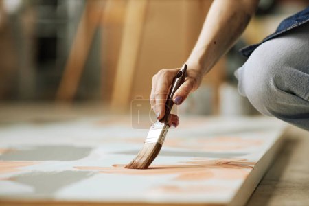 Foto de Enfoque selectivo en la mano de una joven creativa con pincel creando pintura abstracta sobre lienzo mientras se agacha en el suelo - Imagen libre de derechos
